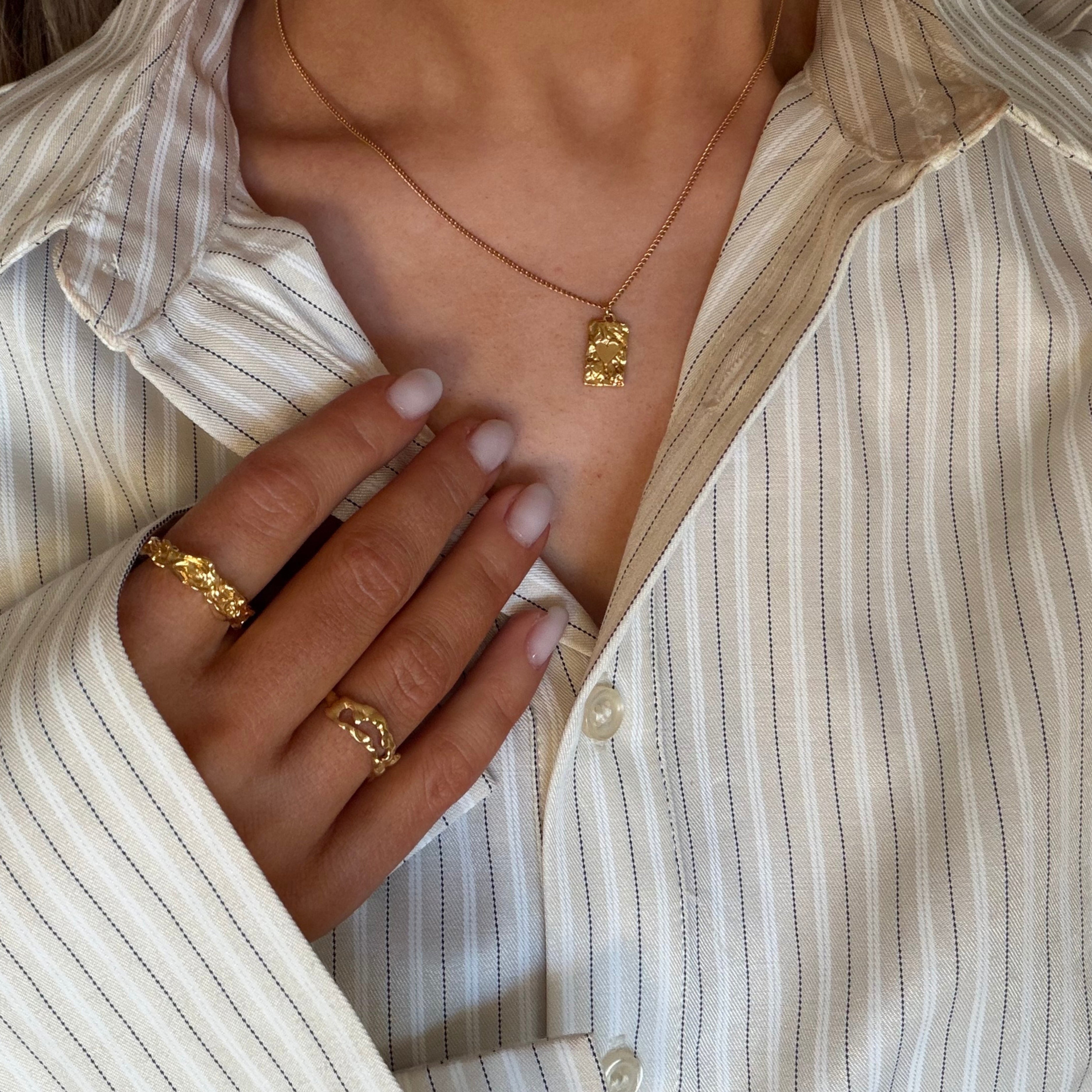 En närbild i livsstilsstil av en person som bär guldsmycken med strukturerad yta – däribland ett halsband med ett litet, strukturerat guldhänge och två strukturerade guldringar – och en skjorta med kritstrecksränder i gräddvitt och brunt.