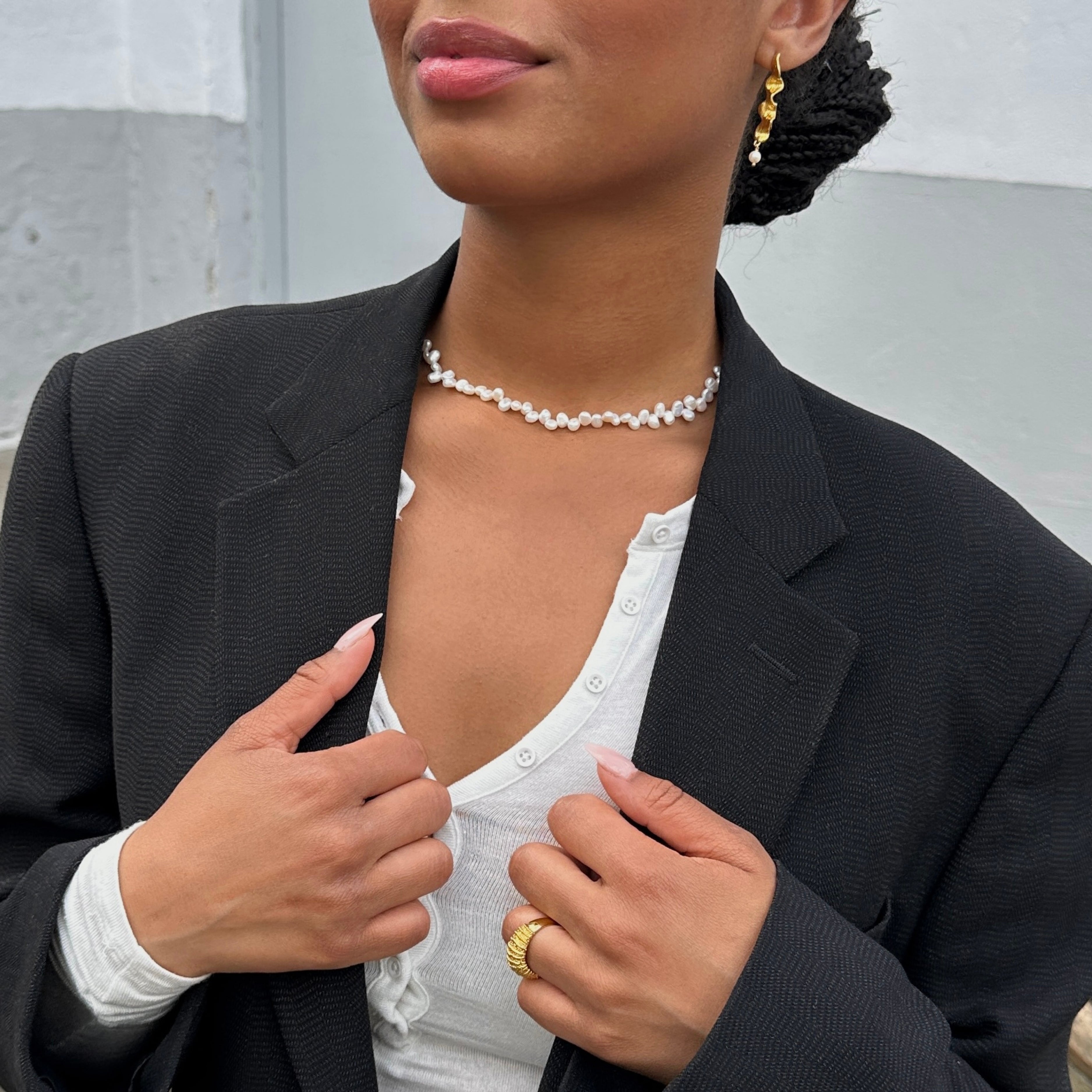 En närbild i livsstilsstil av en modell som bär ett nätt halsband (choker) med vita pärlor, stylat med en svart kavaj och en vit topp.