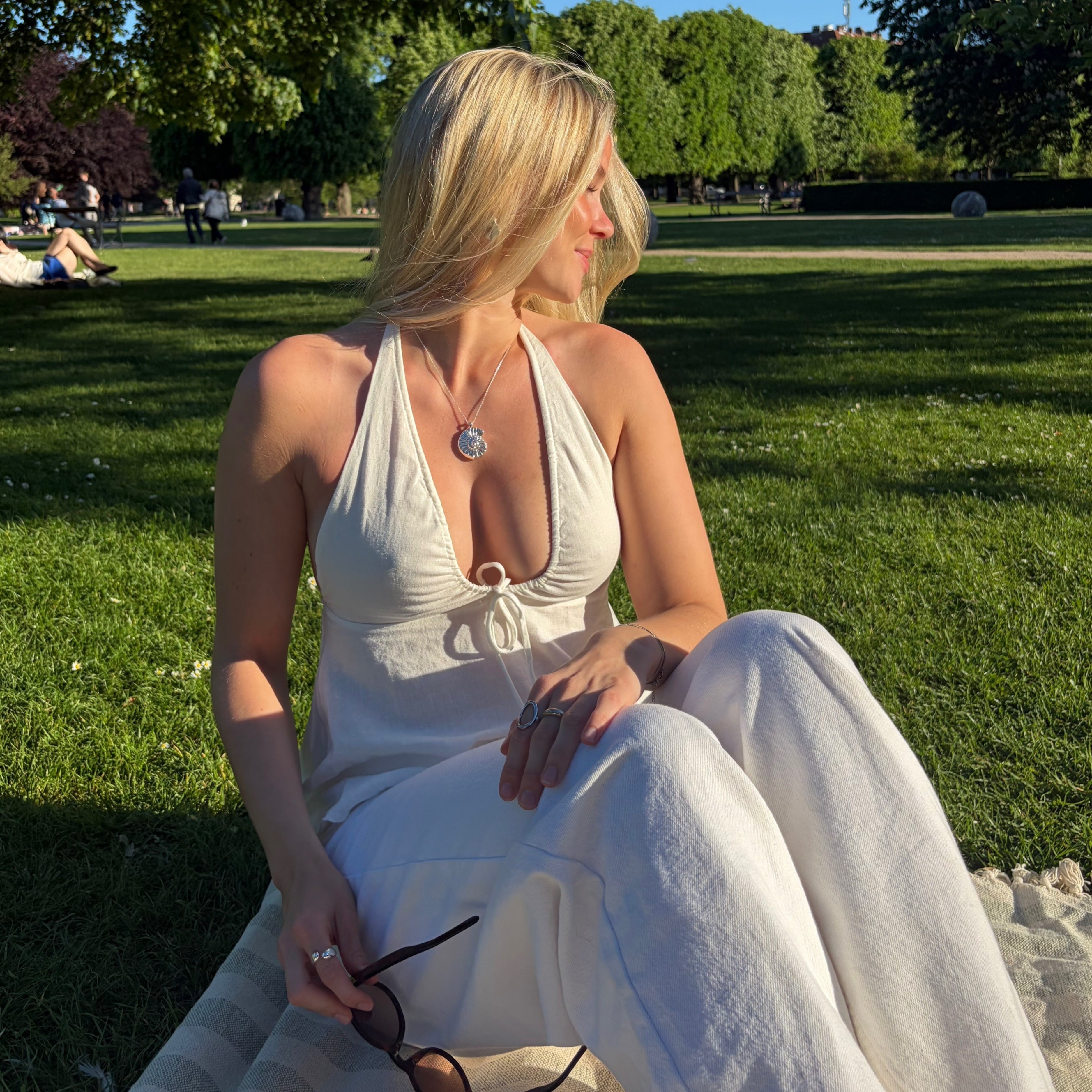 En blond kvinna i vita sommarkläder sitter på en filt i en solig park med gröna träd och gräs i bakgrunden. Hon bär ett silverhalsband med ett snäckskalshänge, håller ett par solglasögon i handen och tittar åt sidan.