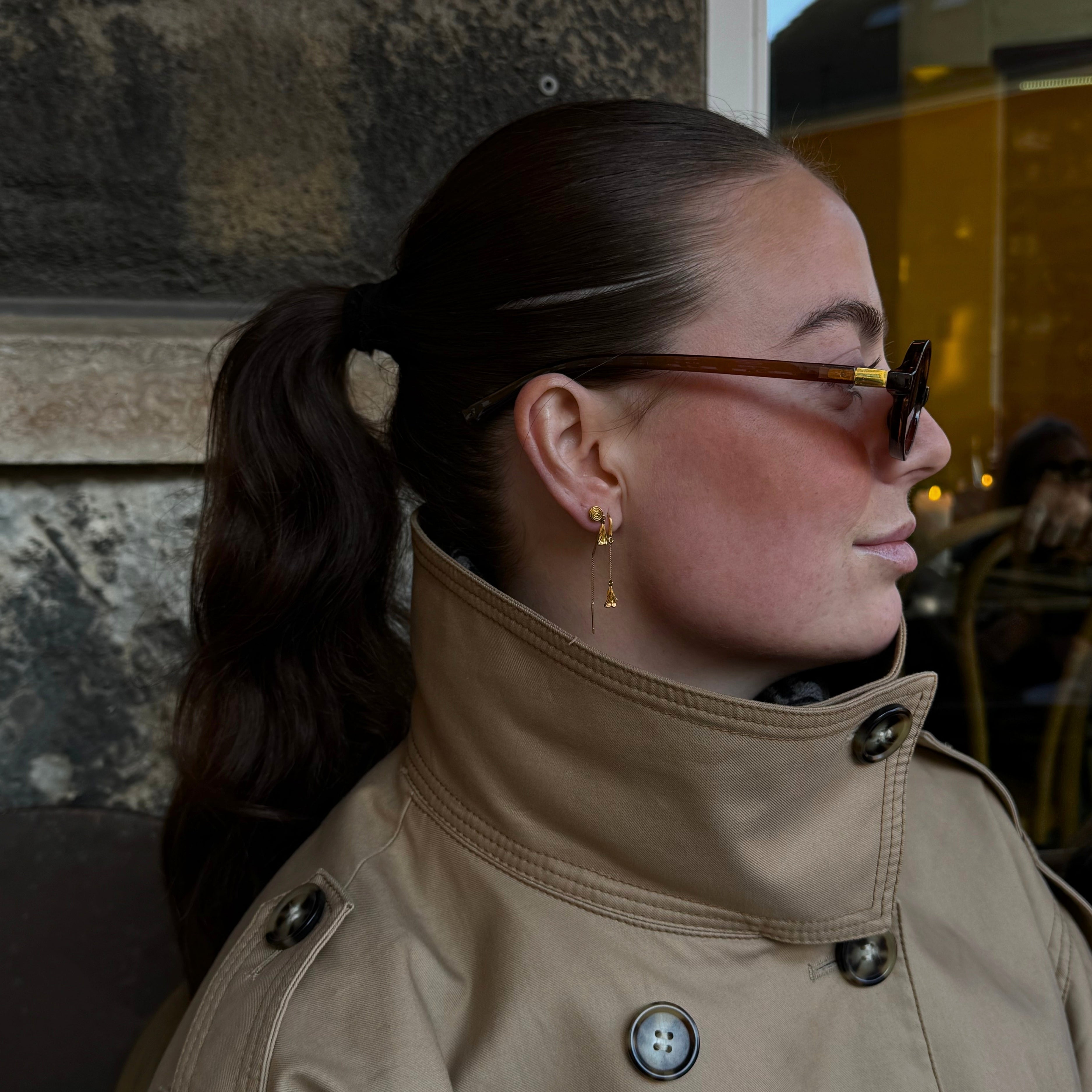En kvinna i profil, iklädd en beige trenchcoat, långa guldörhängen och mörkbruna solglasögon med guldbågar, mot en bakgrund av en stenmur och ett fönster.
