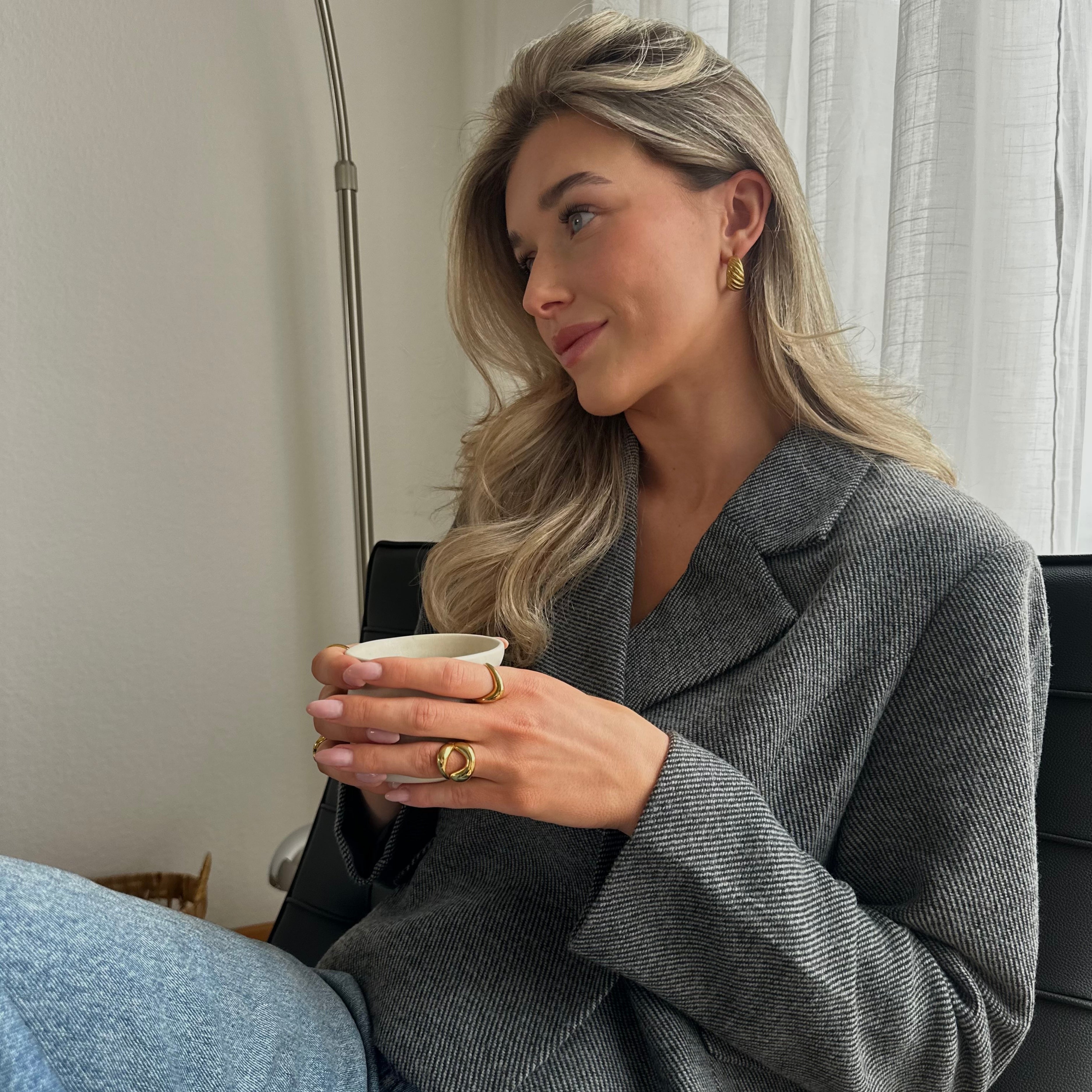 En blond kvinna i grå kavaj och blå jeans håller i en liten vit kopp; hon bär guldringar och ett guldörhänge i en inomhusmiljö med mjuk belysning.