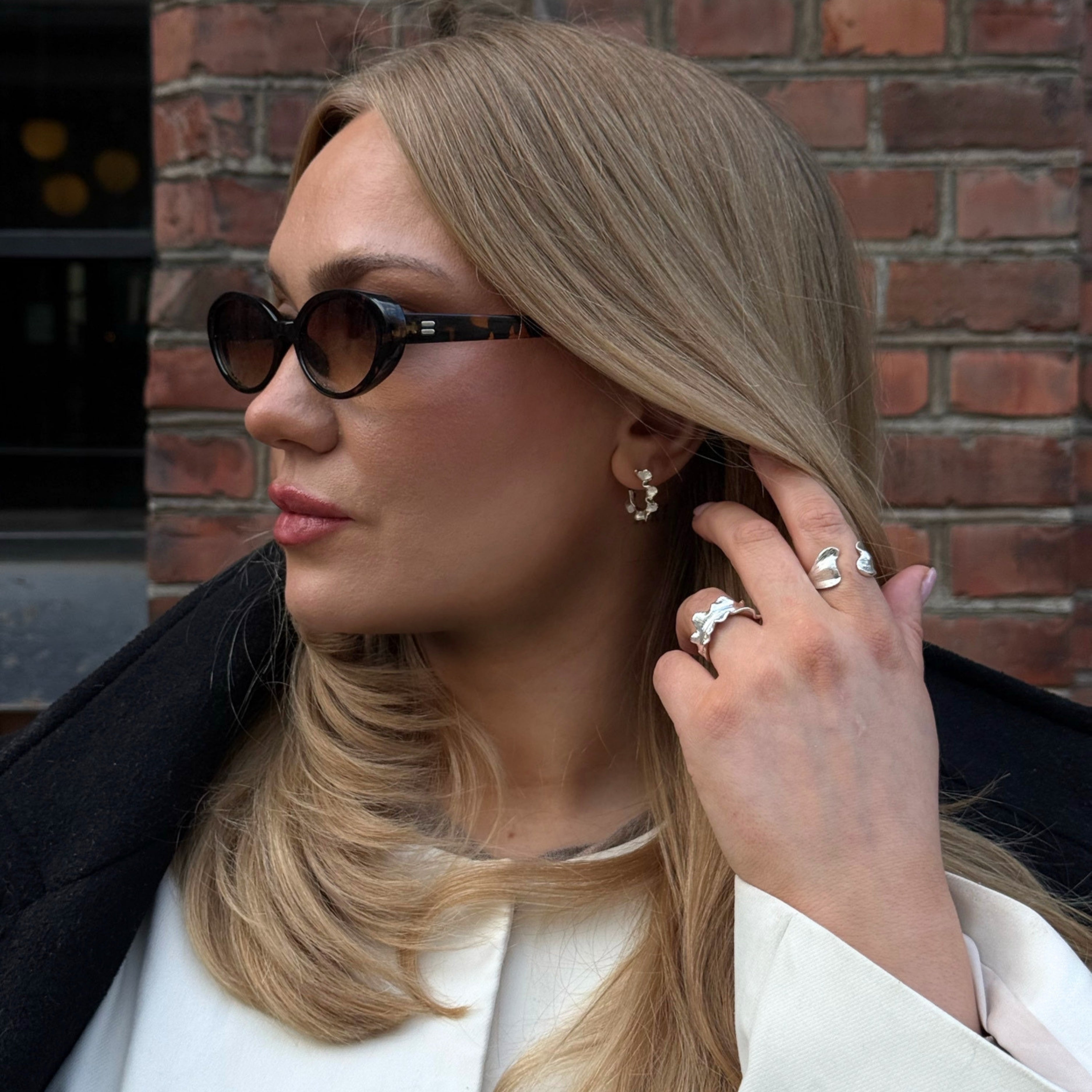 En blond kvinna med kattögonformade solglasögon, stora silverringar i öronen och flera silverringar på fingrarna, som poserar framför en tegelvägg.