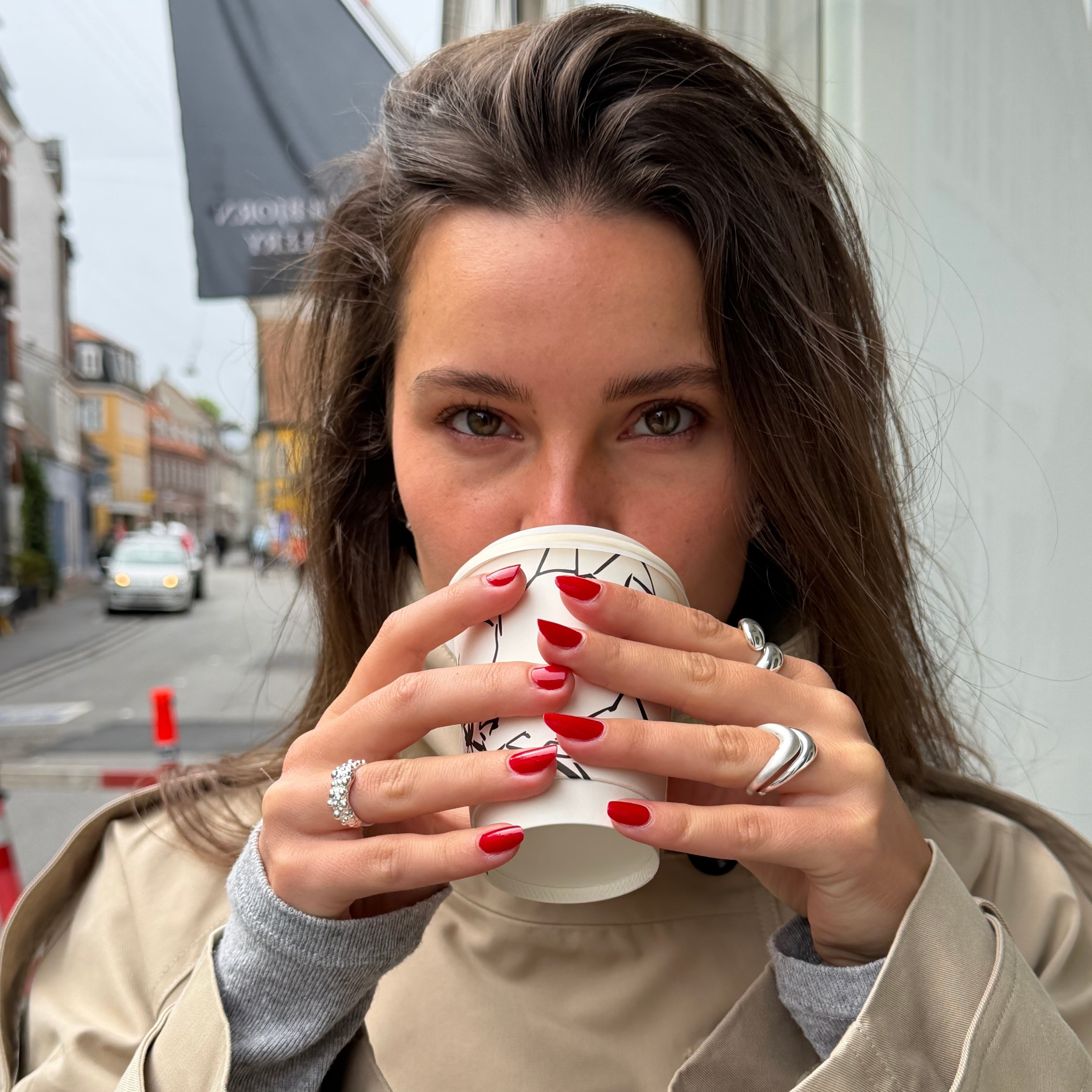 En kvinna med långt, brunt hår håller en vit kaffemugg av papper mot läpparna. Hon bär en beige trenchcoat över en grå överdel och har rött nagellack samt silverringar på sig.