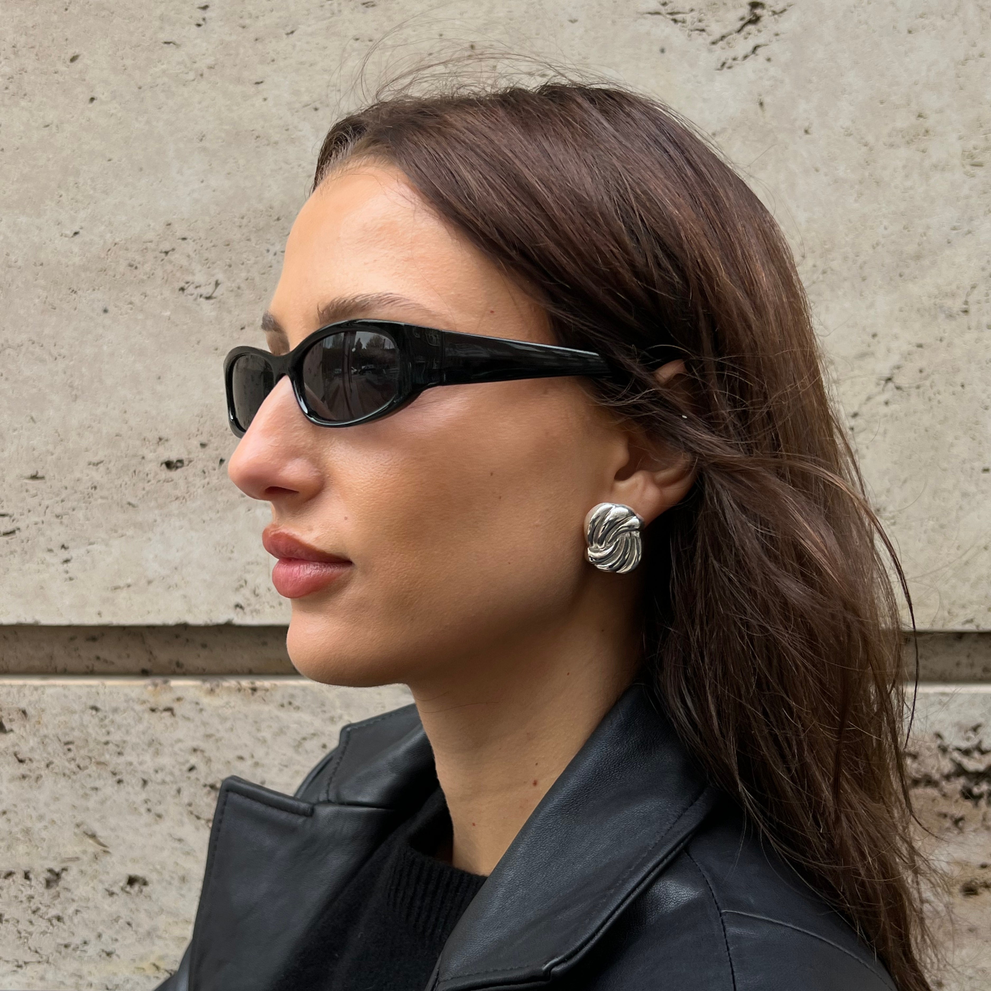 En kvinna i profil, iklädd svarta solglasögon av cat-eye-modell och ett silverfärgat spiralörhänge med struktur, fotograferad mot en neutral betongvägg.